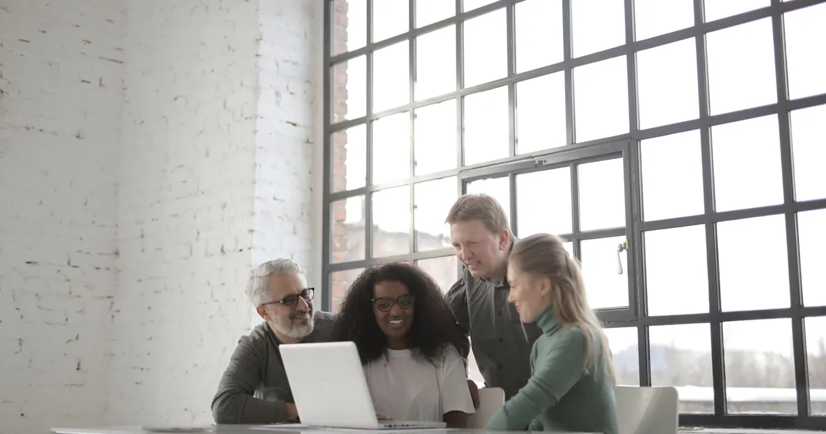 Four colleagues gather around a laptop at a white table in a bright office with tall factory windows.